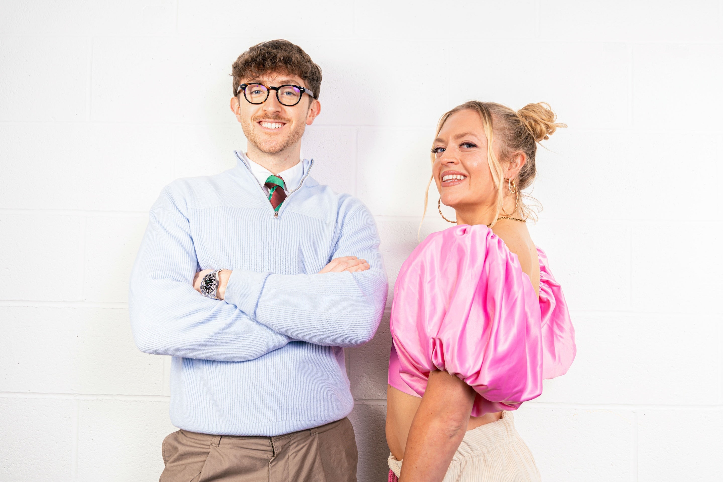Ollie and Bella, Art Dealers in Bristol, Bath and London standing together against a white art gallery wall wearing baby pink silk and a light blue quarterzip. Ollie is wearing a Rolex GMT Master II with black ceramic bezel. 