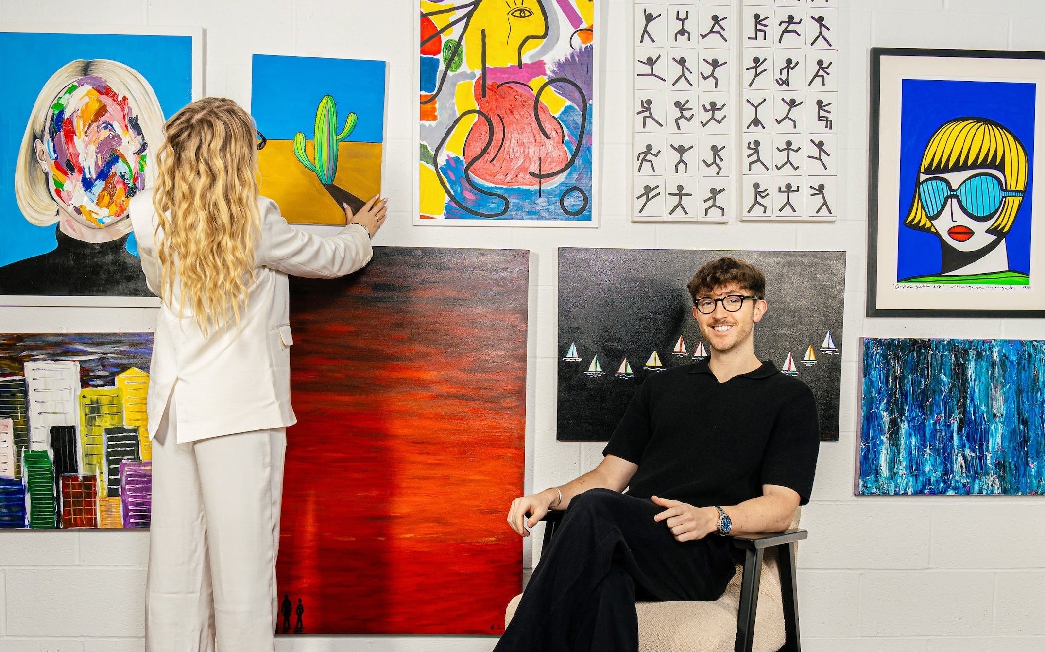 Bella and Ollie art curators standing in front of a colourful abstract art curation on an art gallery wall in Bath, Bristol and London. Bella is wearing Jimmy Fairly glasses and Ollie is wearing Oliver Peoples glasses. Works include Margo in Margate limited edition print. 