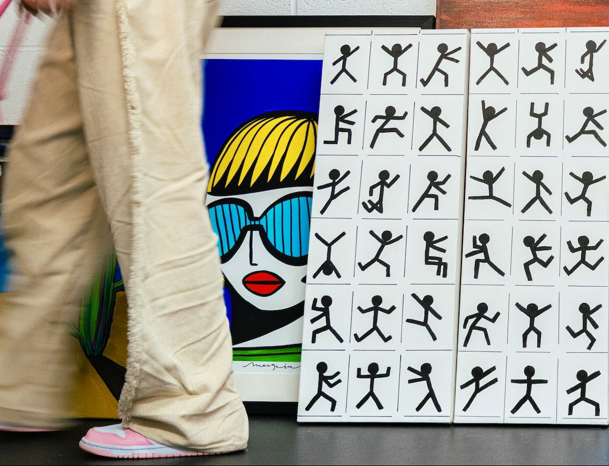 Bella walking past a contemporary, figurative and abstract art curation featuring a limited edition print by Margo in Margate Artist next to a black and white diptyque art piece showing black and white dancing stick men. All artworks are positioned on the floor of the gallery floor. 