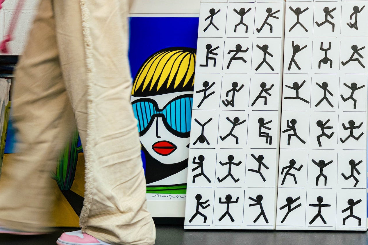 Bella, Art dealer and Art Gallerist, walking past a curation of contemporary, figurative and abstract art on the gallery floor. Girl in the yellow bob, limited edition print by artist Margo in Margate next to figurative black and white stick men art. 