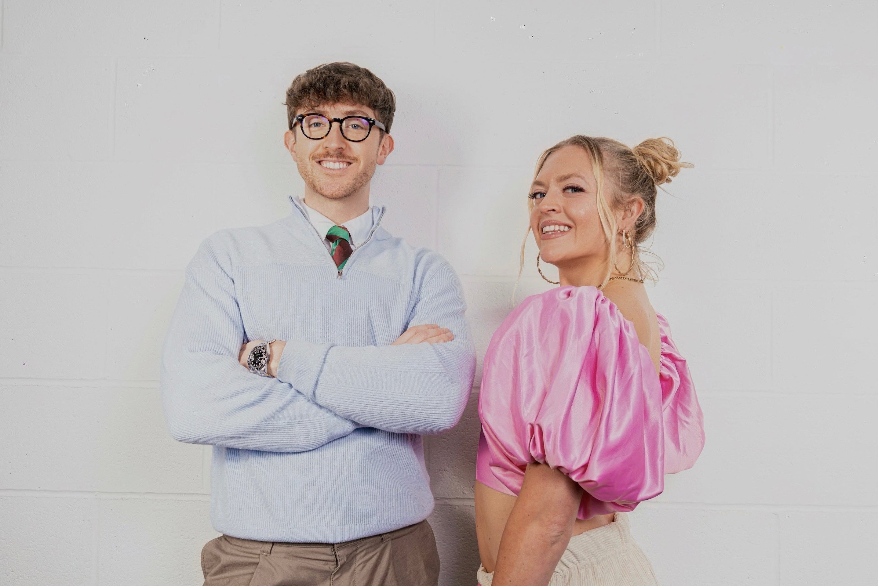 Two people standing against a white tiled wall, one wearing a light blue sweater and the other in a pink top.
