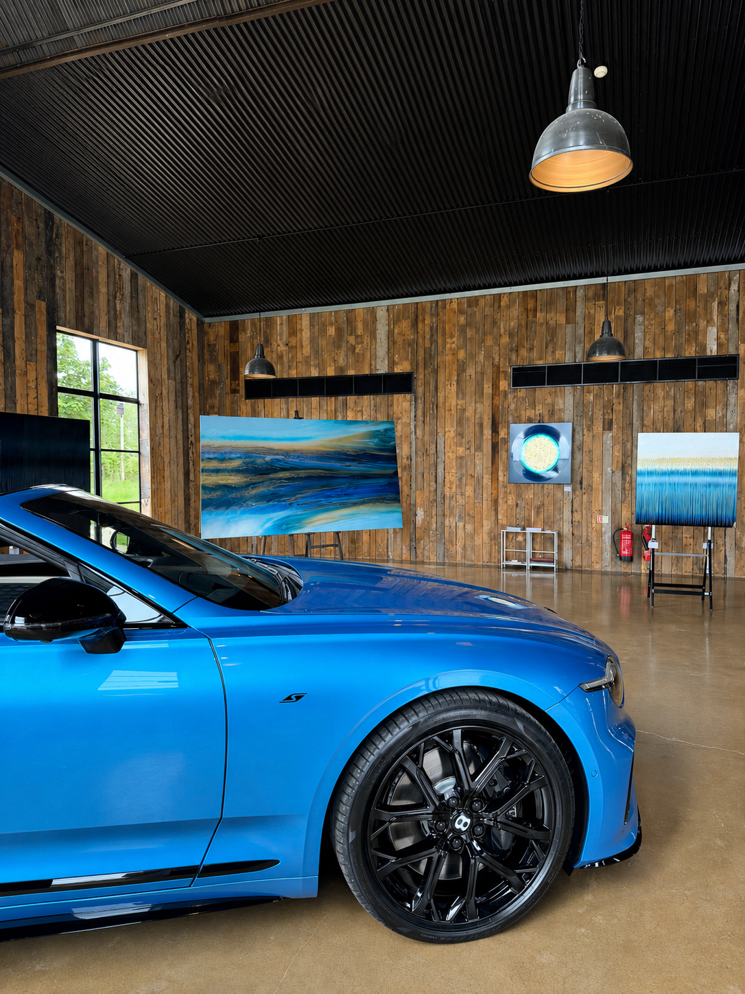Blue Bentley  in a showroom with wooden walls and large windows and artwork by Russell hatton. 