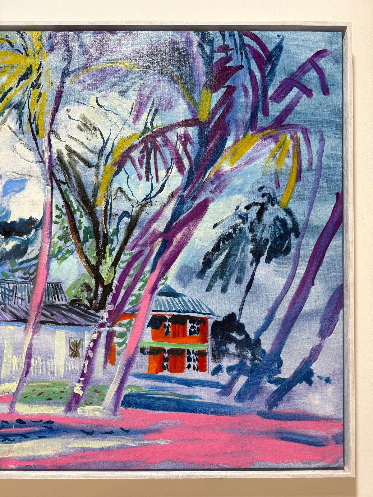 A framed abstract landscape painting featuring stylized palm trees, a small red-orange house, and a pink ground area against a blue and purple sky, shown with a light wood frame. 
Life Beneath the Palms 2026 by Hannah Rollings, original colourful landscape painting inspired by Barbados
