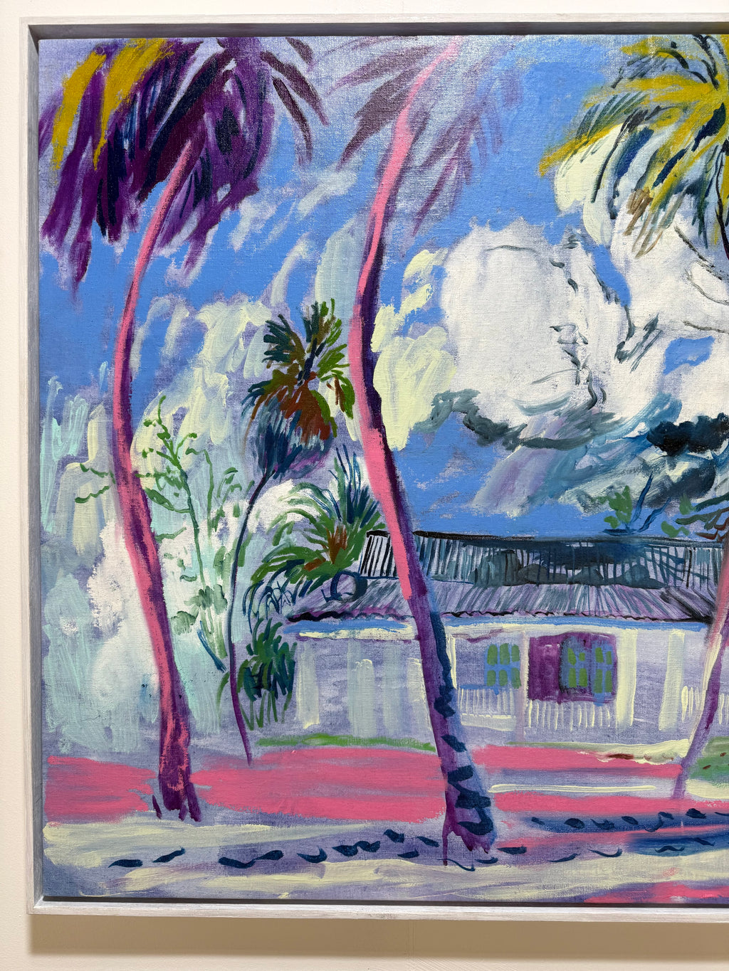 An expressionist-style painting of a tropical beach scene with tall palm trees, a white house, blue sky, and pink ground, shown in a light wood frame. Colourful landscape original art by hannah rollings.