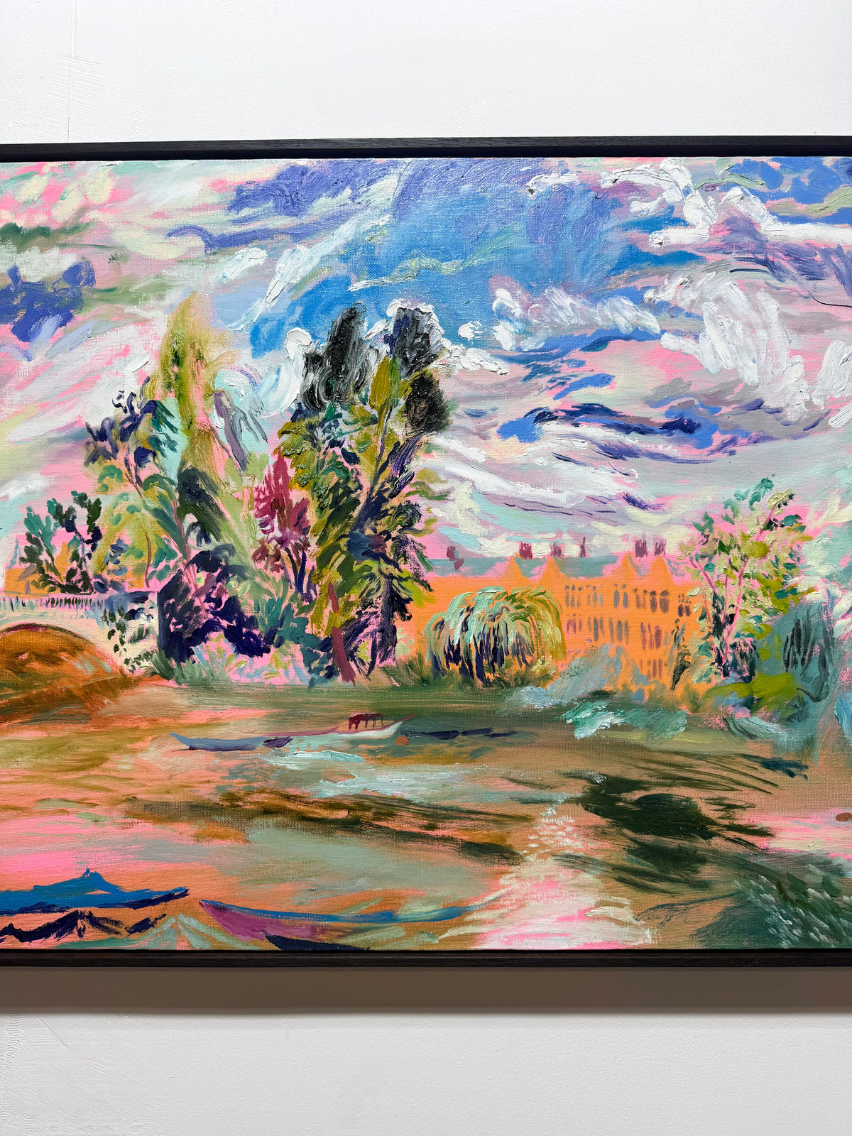 A colorful landscape painting hanging on a white wall, featuring a vibrant sky with blue, pink, and white clouds, green and purple trees, and a warm-toned building reflected in water. The River Carries the Light 2026 by Hannah Rollings, original contemporary landscape painting of the River Thames