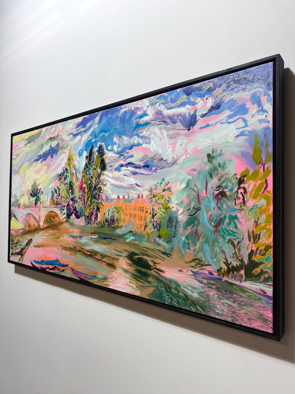A framed horizontal landscape painting with a colorful, impressionistic style, hanging on a plain white wall. The artwork features a bridge, trees, a pinkish path, and a bright sky in shades of blue, pink, green, yellow, and orange, enclosed in a black frame. The River Carries the Light 2026 by Hannah Rollings, original contemporary landscape painting of the River Thames