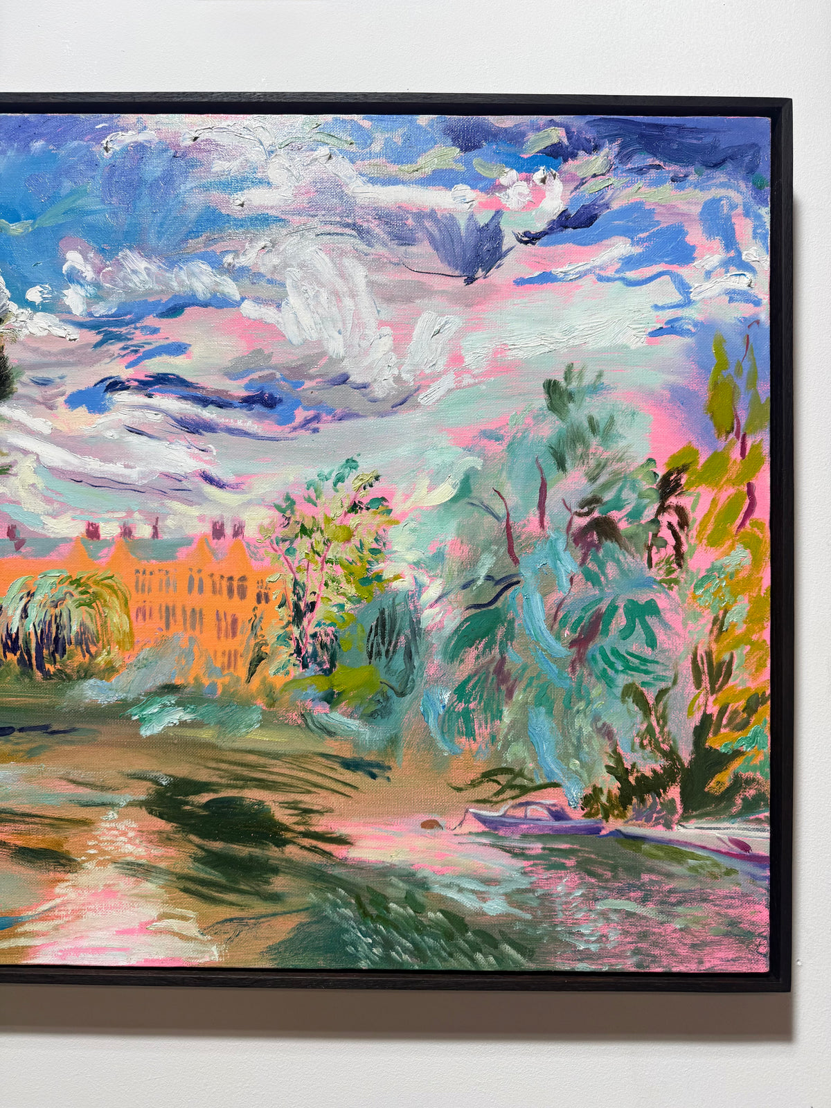 An expressive landscape painting in a black frame, featuring a vibrant sky with swirling blue, pink, and white clouds, green trees, and a warm orange building in the background, hung on a light wall. The River Carries the Light 2026 by Hannah Rollings, original contemporary english landscape painting of the River Thames