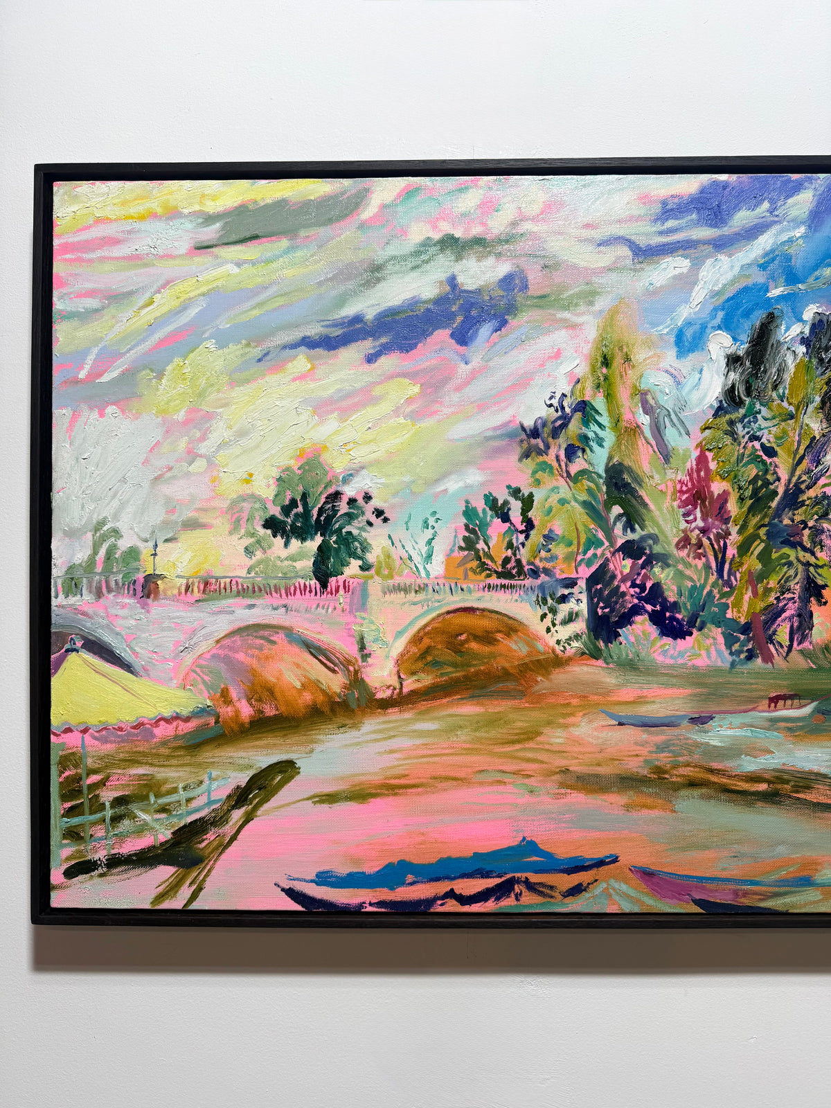 A framed colorful landscape painting on a white wall, featuring expressive brushstrokes in pink, blue, green, yellow, and brown with trees, a bridge, and water. The River Carries the Light 2026 by Hannah Rollings, original contemporary landscape painting of the River Thames